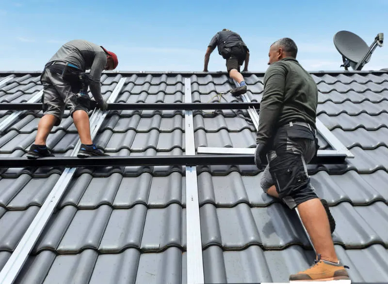 Drei Handwerker auf einem Dach installieren Solarpanels. Sie tragen Arbeitskleidung und Sicherheitsausrüstung und arbeiten an den Dachziegeln, während ein Satellitenschüssel im Hintergrund sichtbar ist.