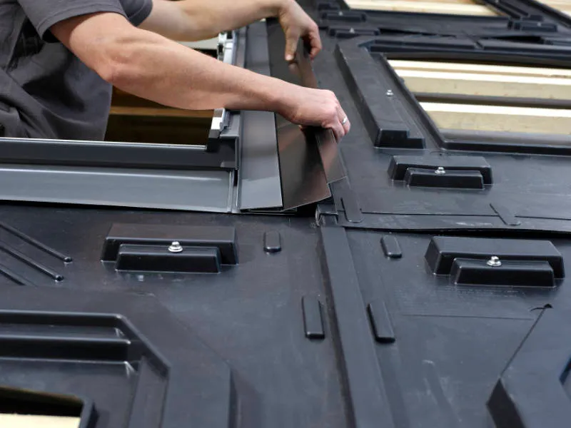 Handwerker montiert ein Dichtungsprofil auf einem Dach, um Solarpanels zu installieren. Die Aufnahme zeigt die Hände des Arbeiters, der an der präzisen Ausrichtung des Materials arbeitet.