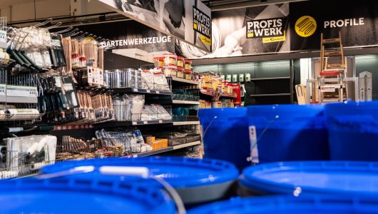 Innenansicht eines Baumarkts mit Regalen voller Werkzeuge und Gartenutensilien von "Profis am Werk". Im Vordergrund sind blaue Eimer sichtbar, während das Markenschild deutlich im oberen Bereich des Bildes zu sehen ist.