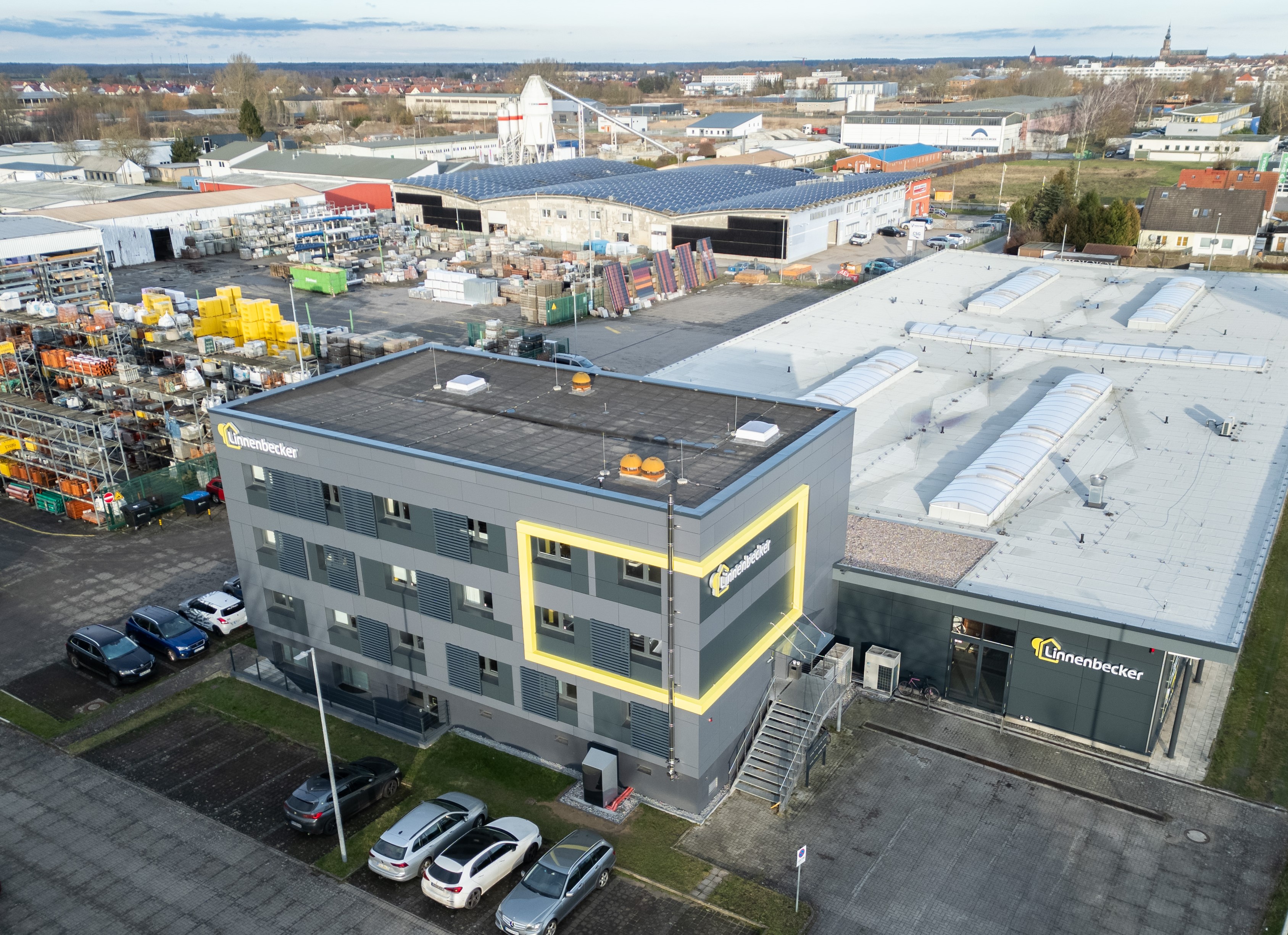 Luftaufnahme eines modernen Bürogebäudes mit gelber Akzentfarbe, das von einem größeren Industriekomplex umgeben ist. Auf dem Parkplatz sind mehrere Autos zu sehen, und im Hintergrund sind weitere Fabrikhallen sichtbar.