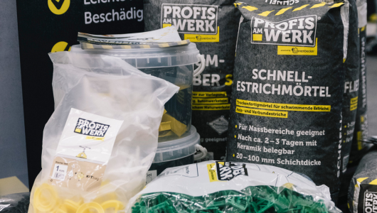 Baustellenmaterialien, einschließlich Säcken mit "Schnell Estrichmörtel", Kunststoffkeilen und weiteren Produkten von "Profis am Werk". Die Produkte sind auf einem Tisch oder Regalen angeordnet, mit klar sichtbarem Branding.