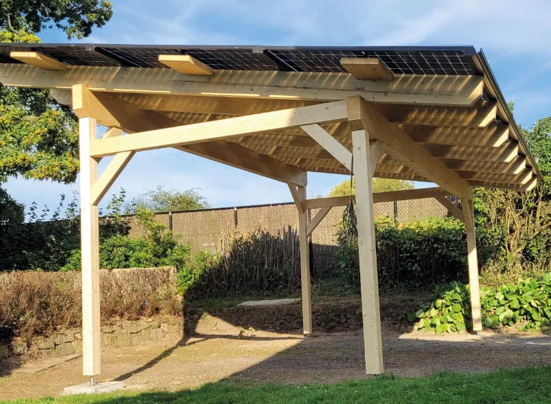 Freistehende Holzstruktur mit Solarpanels auf dem Dach, die in einem Garten installiert ist. Der Pavillon bietet Schutz vor Sonne und Regen und ist von grüner Vegetation umgeben.