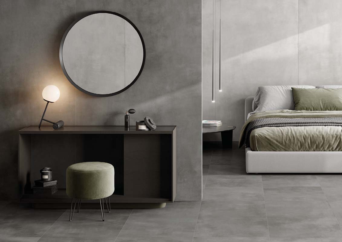 Modernes Schlafzimmer mit einem minimalistischen Design, grauen Wänden und Bodenfliesen. Ein runder Spiegel, ein grüner Hocker und eine stilvolle Lampe ergänzen den Raum. Das Bett hat Bettwäsche in sanften Grüntönen.