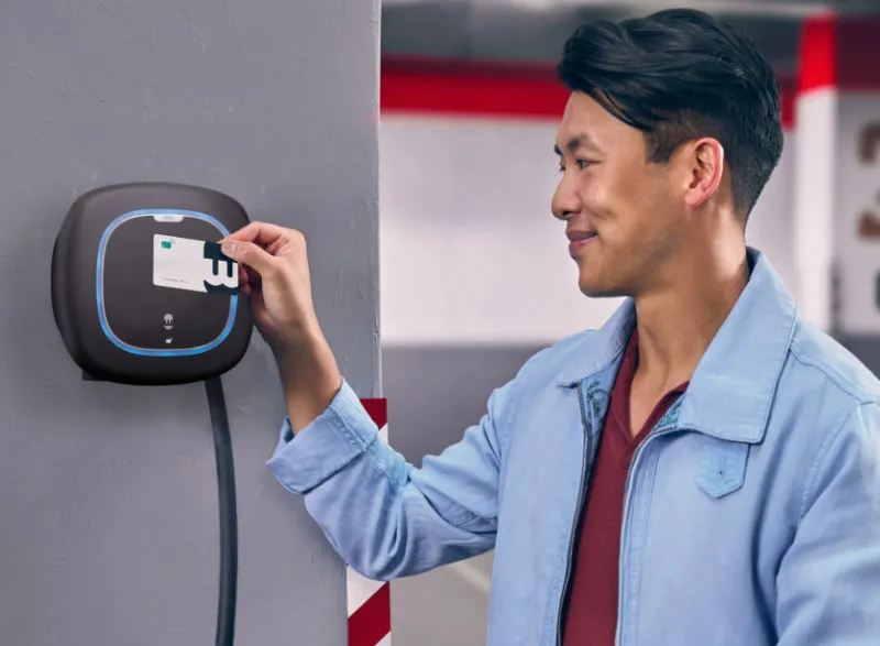 Mann nutzt eine RFID-Karte, um eine Ladestation für Elektroautos in einer Tiefgarage zu aktivieren. Die Ladestation ist an der Wand montiert und leuchtet blau.