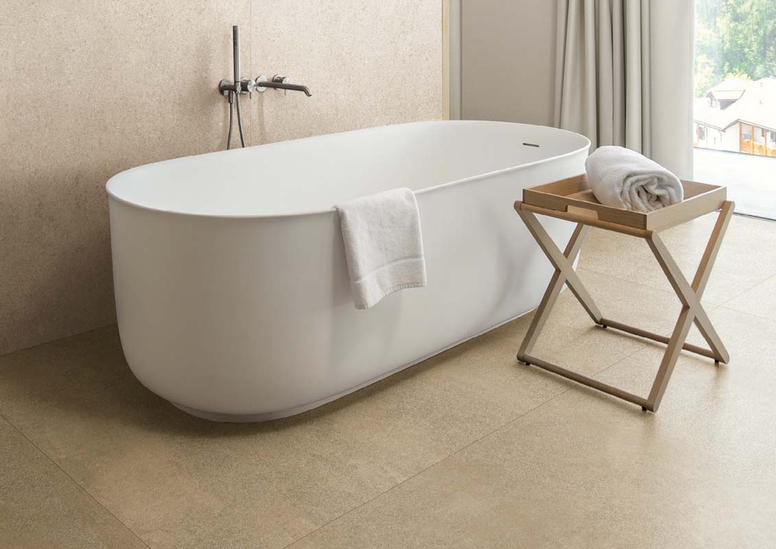 Elegante freistehende Badewanne mit einem weißen Handtuch, ein Beistelltisch aus Holz mit einem weiteren Handtuch und beige Fliesen im Badezimmer. Große Fenster bieten einen Blick auf die Natur.