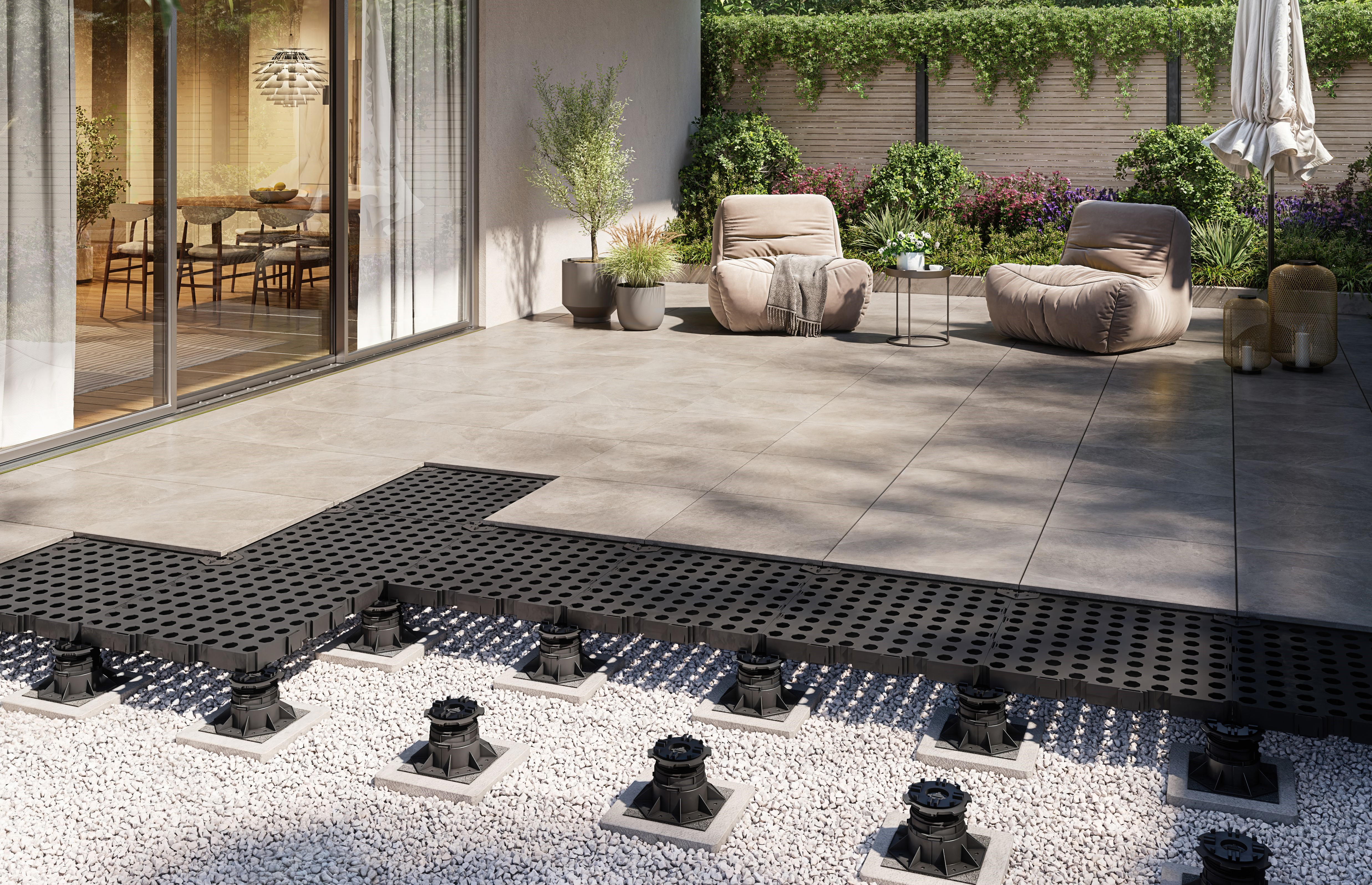 Modernes Terrassendesign mit großen Fliesen, einer grauen Steingartenfläche und verstellbaren Unterkonstruktionen. Zwei bequeme Sessel und ein Tisch stehen auf der Terrasse, die von grünen Pflanzen umgeben ist.