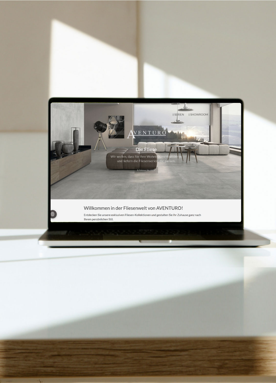 Laptop auf einem Tisch, der eine Webseite mit dem Titel "Willkommen in der Fliesenwelt von AVENTURO" anzeigt. Im Hintergrund ist ein modernes Wohnzimmer mit großen Fliesen und Möbeln sichtbar.