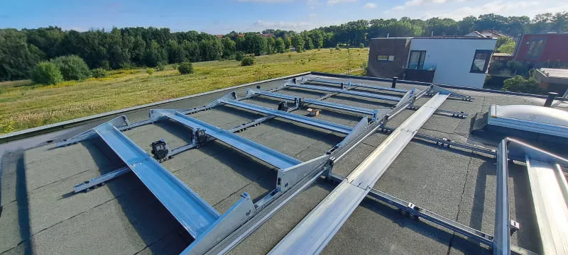 Blick auf ein flaches Dach mit installierten Montagestrukturen für Solarpanels. Im Hintergrund erstreckt sich eine grüne Landschaft, die das Dach und die umliegenden Gebäude überblickt.