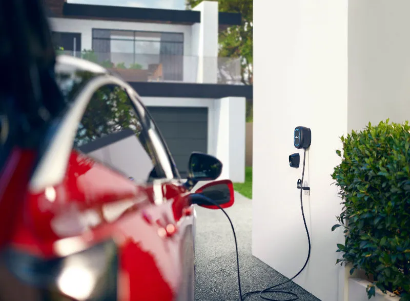 Roter Elektroauto wird an einer Wandladestation aufgeladen, die an einem modernen Gebäude installiert ist. Das Kabel ist mit dem Auto verbunden, während die Ladeeinheit an der Wand des Hauses befestigt ist.