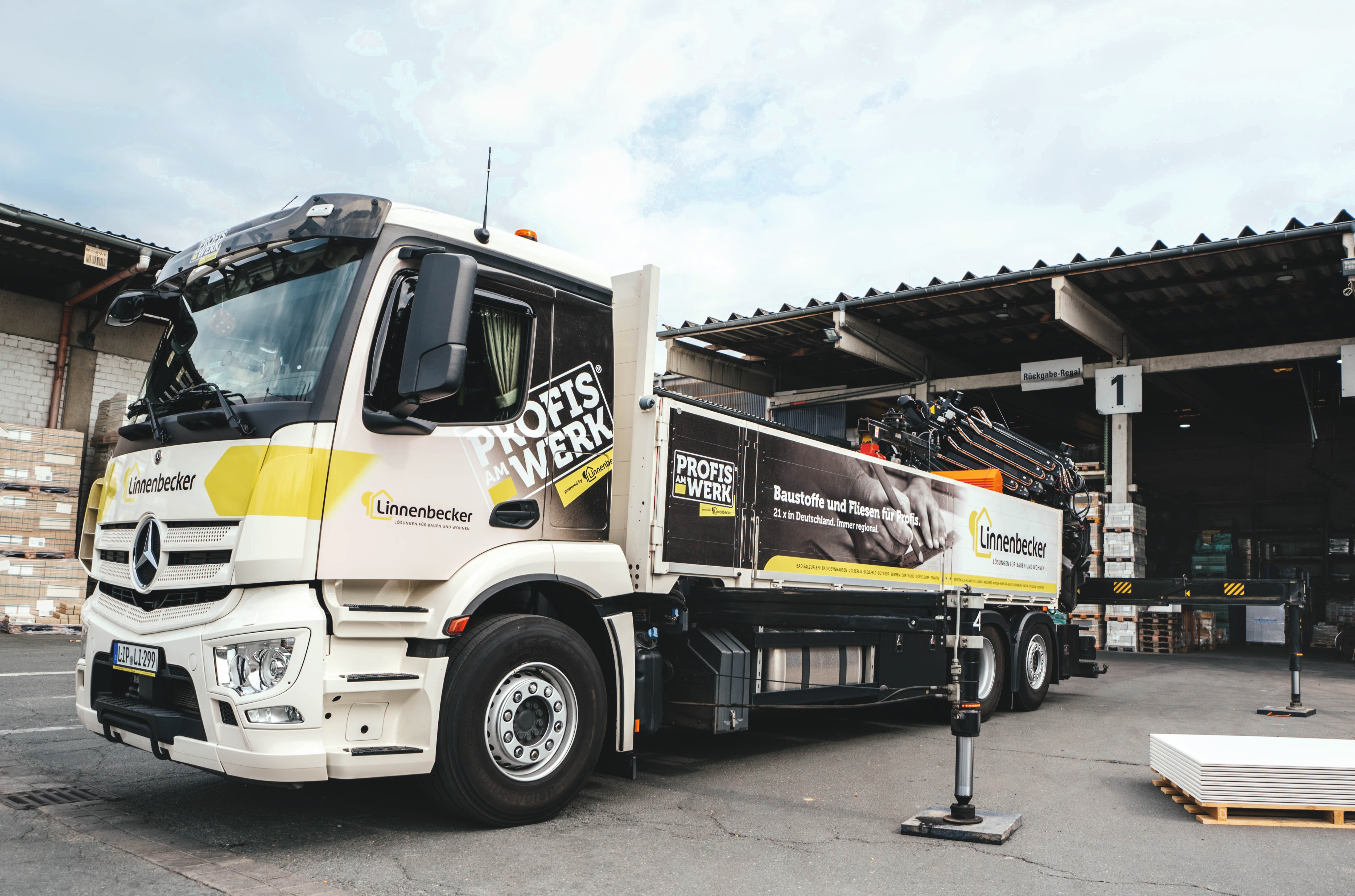 Lkw der Marke Mercedes-Benz mit einem Kranaufbau und dem Logo "ProfisWerk" auf der Seite, parkt vor einem Lagerhaus. Der Lkw ist mit gelben und schwarzen Akzenten versehen und in einem industriellen Umfeld positioniert.